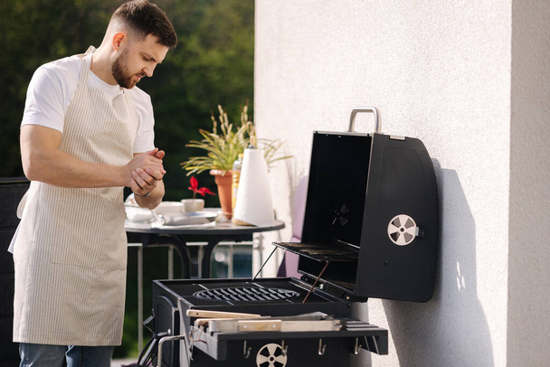 Side view of handsome man try how hot is BBQ grill. Man in apron make dinner outdoor. High quality photo - Photo, Image
