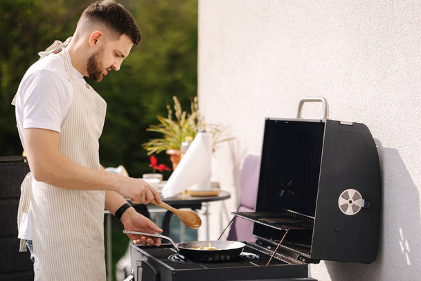 Красивый мужчина готовит на открытом воздухе на гриле. Человек жарит еду на сковородке. Летняя еда на свежем воздухе. Высокое качество фото - Фото, изображение