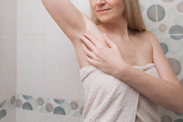 Positive beautiful woman with blond hair and in a white towel, touches her shaved armpits in the bathroom. Image on the theme of health, hygiene and personal care. - Foto, immagini