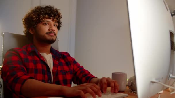Um homem com cabelo encaracolado e barba olha para algo no monitor e digita no teclado. Um jovem com uma camisola xadrez trabalha em um computador de seu escritório em casa. Imagens 4k de alta qualidade - Filmagem, Vídeo