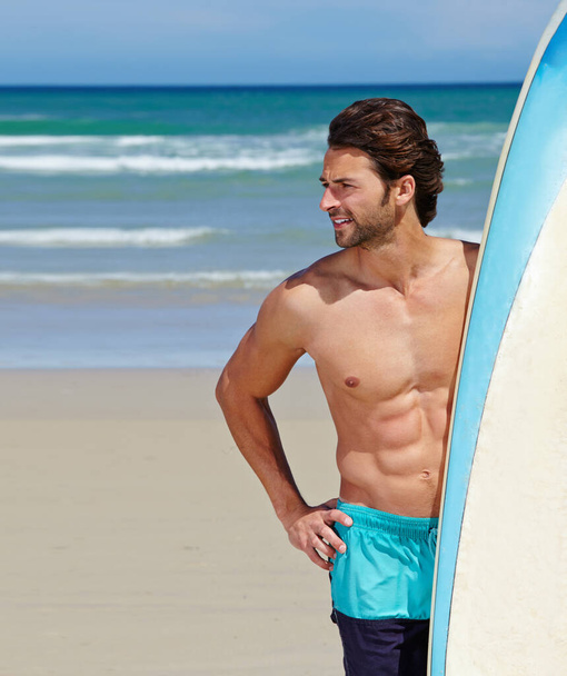 A day at the beach. A young man standing next to his surfboard looking around - Photo, Image