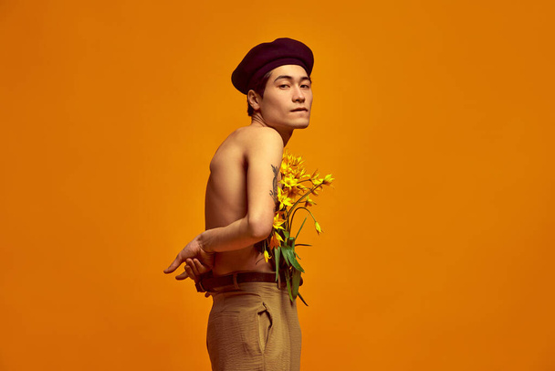 Portrait of stylish, young, korean guy, model posing shirtless in beige pants, with sticking out flowers against yellow studio background. Concept of mens fashion, style, art, beauty, trends - Fotó, kép
