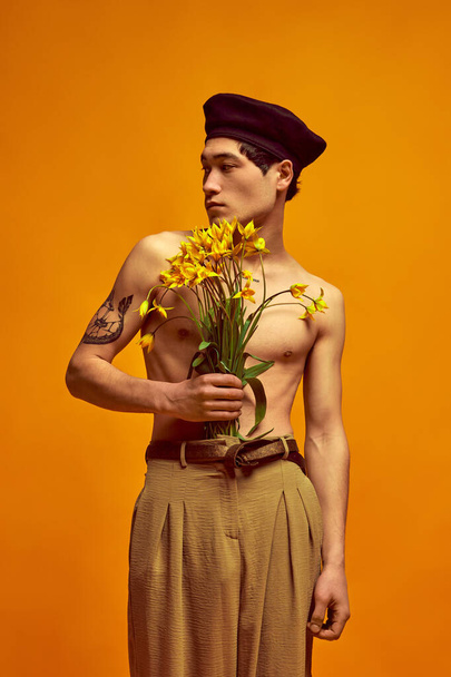 Parisian spring and summer collection. Handsome korean guy posing shirtless in classical pants, holding flowers against yellow studio background. Concept of mens fashion, style, art, beauty, trends - Photo, Image
