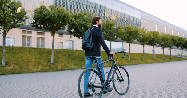 Back view of attractive calm modern tall young bearded man in jacket and jeans which walking with his bike on cobbled street near beautiful modern city building - Zdjęcie, obraz
