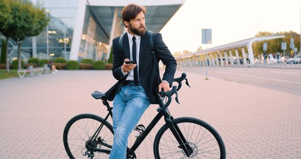Handsome confident positive young bearded man standing near his bike using phone on the city airport background - Foto, imagen