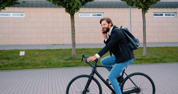 Likable confident purposeful 30-aged modern businessman with well-groomed beard in black jacket and jeans has mobile conversation while going to his job on bicycle on city cobbled street,slow motion - Фото, изображение