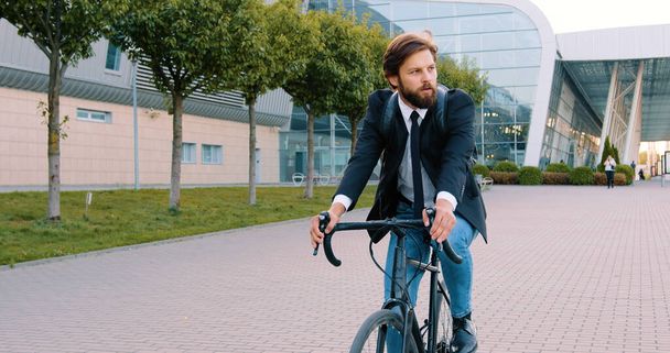 Приятный вдумчивый молодой бородатый бизнесмен с рюкзаком в формальной одежде едет на велосипеде в свой офис на мощеной улице рядом с большим панорамным зданием - Фото, изображение