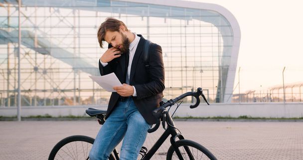 Good-looking serious busy purposeful 30-aged bearded businessman in formal clothes leaning on his bicycle while discussing financial papers with colleague on smartphone during stop near modern urban - Fotoğraf, Görsel