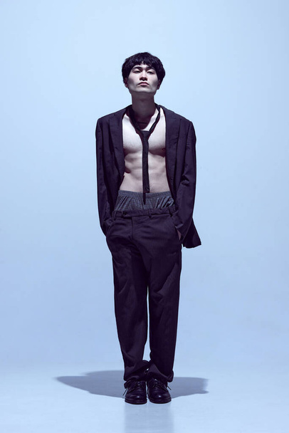 Portrait of young stylish man posing in black jacket on naked body with tie and classical pants against light blue studio background. Concept of mens fashion, style, art, beauty, trends - Fotó, kép