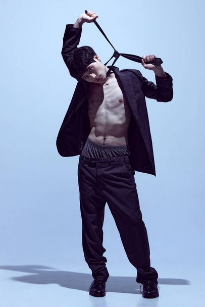 Portrait of stylish young guy posing in black jacket on naked body, tie and classical pants against light blue studio background. Concept of mens fashion, style, art, beauty, trends - 写真・画像