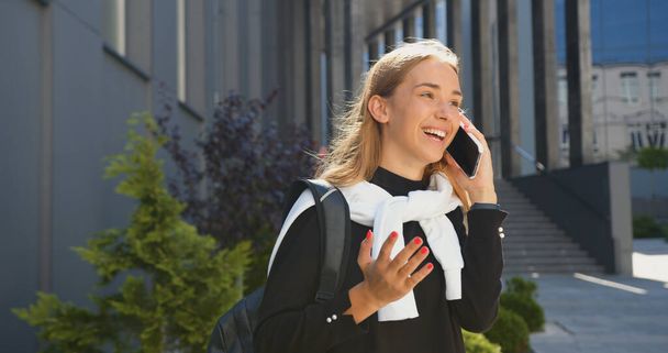 Portrait of good-looking trendy smiling lucky young light-haired girl in casual clothes with backpack which standing near modern city building and talking on smartphone with friend - Fotó, kép