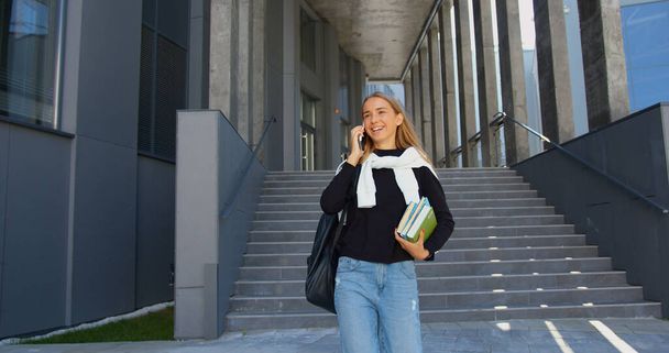 Beautiful smiling satisfied young light-haired girl student in casual clothes going down the steps while leaving university and enjoying her phone conversation - Valokuva, kuva
