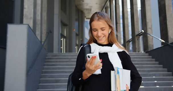 Gorgeous happy joyful young girl with light long hair in casual clothes carrying her books and enjoying music from phone playlist while going down the steps from university or office building - Фото, изображение