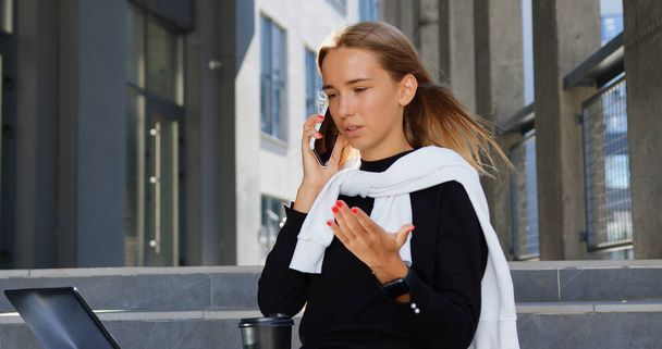 Beautiful concentrated modern young light-haired girl in stylish youthful clothes sitting on buildings stairs and has hard business conversation with worker or subordinate while working on laptop - 写真・画像