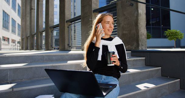 Attractive smiling satisfied young light -haired girl in trendy youthful clothes sitting on buildings steps,working on laptop,drinking coffee from paper cup and talking on phone - 写真・画像