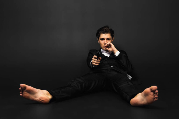 Young mafia member with a slicked back hair, in black suit, smokes a cigar, pointing a gun or pistol at the camera. dangerous Italian mafia gangster. isolated on black. - Photo, Image