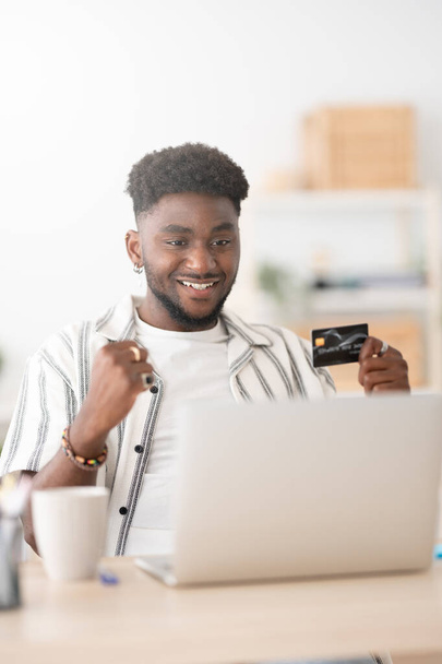 Feliz hombre negro de compras en línea en el ordenador portátil. Afroamericano celebrando compra y tenencia de tarjeta de crédito. - Foto, Imagen