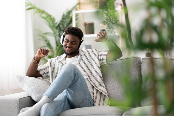 Black young man listening to music and dancing on sofa. - Foto, imagen