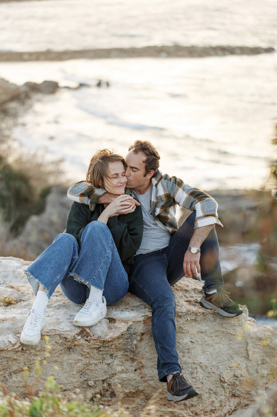 キプロスのビーチで岩の上に座っている中年のカップルの肖像画。彼らはカジュアルな服を着ている。関係概念. - 写真・画像