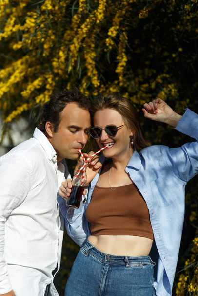 Young happy couple outdoors drinking cold drink from glass bottle with straw. Side view of man and woman drinking soft drink and smiling. Yellow flowering on background. - Zdjęcie, obraz