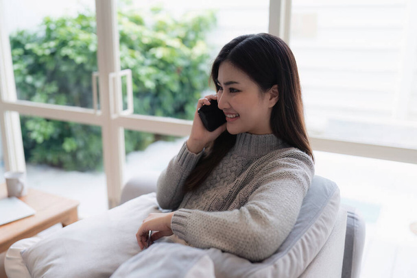 Houkutteleva aasialainen nainen lepää mukava olohuone ja matkapuhelimella, rentoutua, sohva, elämäntapa. - Valokuva, kuva