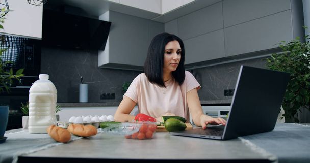 Schöne lächelnde zufriedene junge Brünette sitzt am Küchentisch und schaut während der Zubereitung von Gemüsesalat für das Familienessen das kulinarische Programm online auf dem Laptop - Foto, Bild