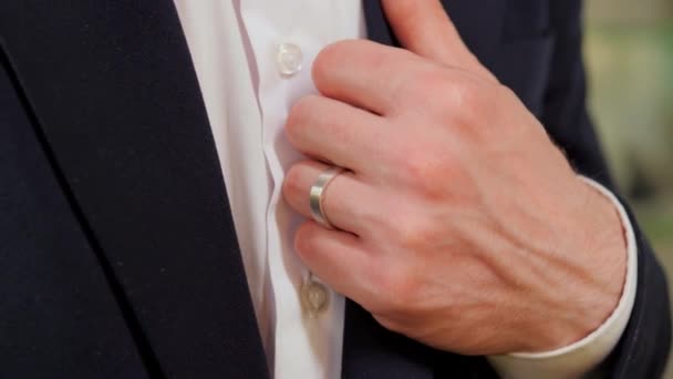 Elegante mano de hombre con un hermoso anillo de oro blanco coloca su mano en el cuello de la chaqueta en vista de cerca. Esta imagen transmite elegancia, estilo y atención al detalle. Tendencia gente de negocios en traje formal - Imágenes, Vídeo