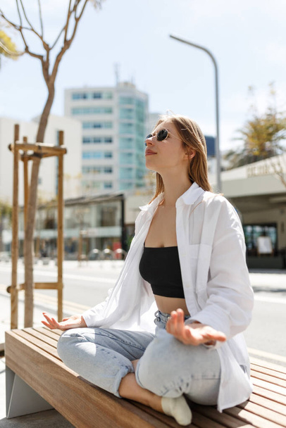 Beautiful young adult woman meditating sitting crossed leg lotus pose in white shirt, blue jeans in Cyprus, Nicosia city. Peaceful break time in busy city life - Zdjęcie, obraz