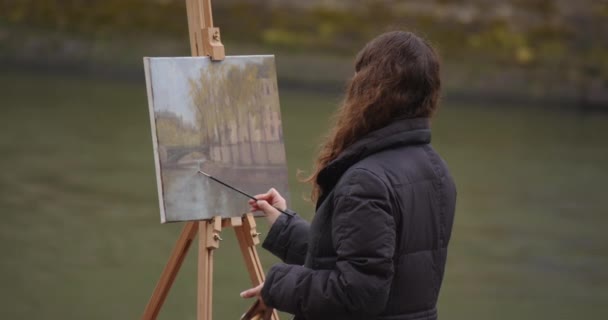 Γυναίκα ζωγραφίζει μια εικόνα κοντά στον ποταμό Σηκουάνα. Δημιουργική γυναίκα καλλιτέχνης ζωγραφίζει μια εικόνα με ένα πινέλο, ενώ στέκεται κοντά στο καβαλέτο στο Παρίσι. Γυναίκα καλλιτέχνης, τέχνη για τις πωλήσεις, έμπνευση. - Πλάνα, βίντεο