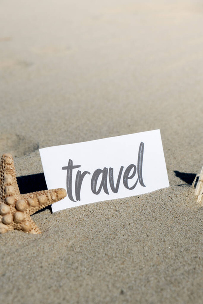 TRAVEL szöveg papíron üdvözlő kártya hátterében tengeri kagyló nyári nyaralás dekoráció. Homokos strand napsütötte part. Ünnepi képeslap. Utazás Üzleti koncepció - Fotó, kép