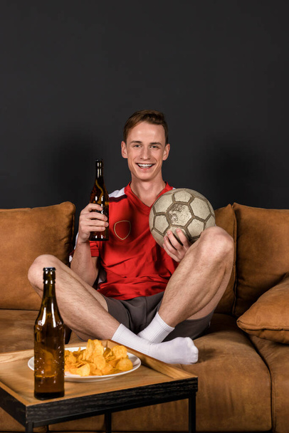 Футбольний фанат у червоній футболці дивиться матч по телевізору, сидить на дивані, п'є пиво і їсть чіпси
. - Фото, зображення