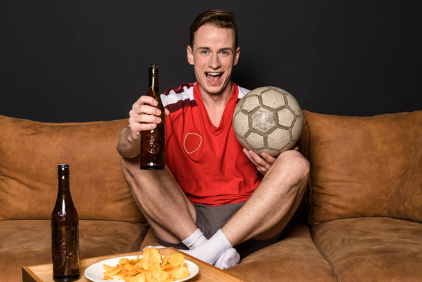 赤いジャージーのサッカーファンは、テレビで試合を見たり、ソファに座ってビールを飲んだり、チップを食べたりしています. - 写真・画像