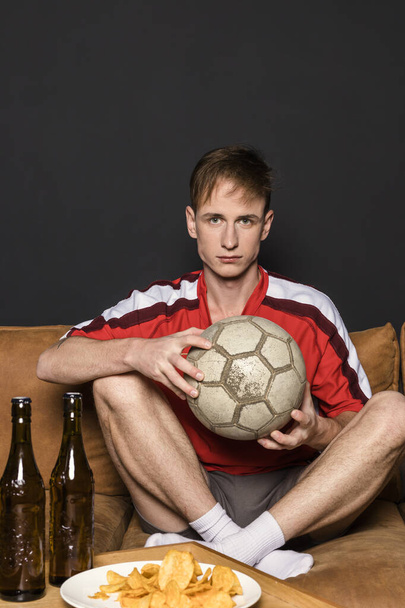 Portrait of a football fan with a ball, in a red jersey is watching a match on TV, sitting on a couch, drinking beer, and eating chips. - Фото, изображение
