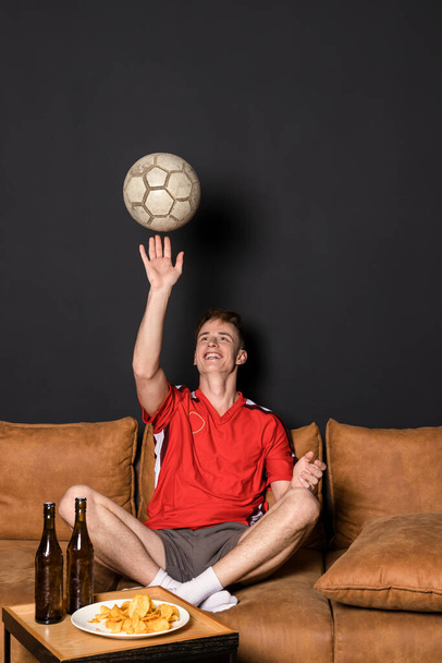 Portret kibica piłki nożnej z piłką, w czerwonej koszulce ogląda mecz w telewizji, siedzi na kanapie, pije piwo i je frytki. - Zdjęcie, obraz