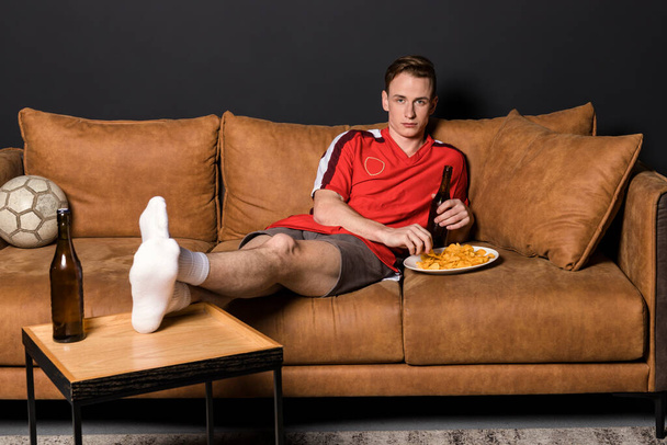 Un fanático del fútbol en una camiseta roja está viendo un partido en la televisión, acostado en un sofá, bebiendo cerveza y comiendo papas fritas.. - Foto, imagen