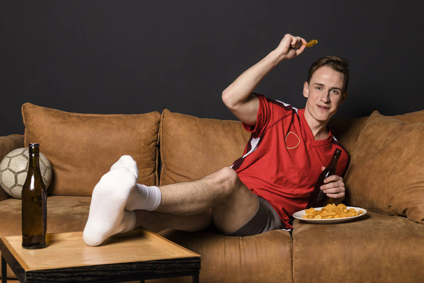 Футбольный фанат в красной майке смотрит матч по телевизору, лежит на диване, пьет пиво и ест чипсы. - Фото, изображение