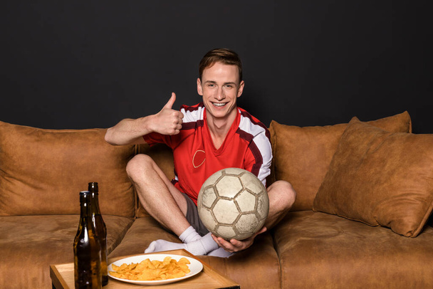 Um fã de futebol numa camisola vermelha está a ver um jogo na TV, sentado num sofá, a beber cerveja e a comer batatas fritas.. - Foto, Imagem