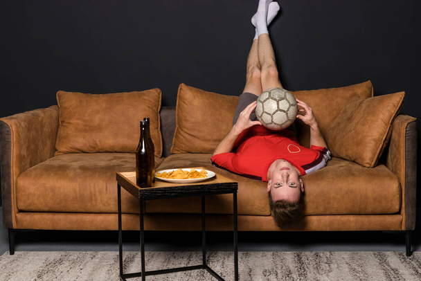 Fotbalový fanoušek Nudil se s míčem, v červeném dresu se dívá na zápas v televizi, sedí na gauči, pije pivo a jí hranolky. Nudný fotbalový zápas. oblíbený tým prohrává. - Fotografie, Obrázek