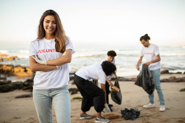 Ευτυχισμένοι νέοι και διαφορετικοί άνθρωποι εθελοντές με μπλουζάκια και Ασιάτισσα με σακούλες σκουπιδιών καθαρίζουν σκουπίδια στην παραλία, σε εξωτερικούς χώρους. Θαλάσσια ρύπανση και προστασία του περιβάλλοντος, προστασία του πλανήτη, Ημέρα της Γης - Φωτογραφία, εικόνα