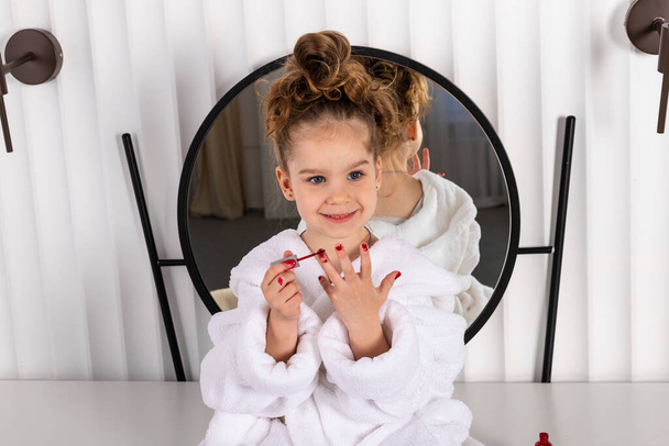 Adorable little girl with a long curly brown hair, in white bathrobe, sitting against a mirror and playing around with her mothers makeup, painting her fingers with nail polish. - Photo, Image
