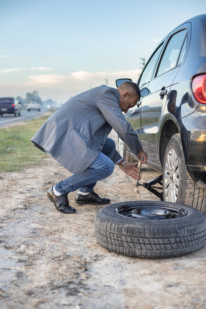 Latino man on suit changes a flat tire on his car at the side of the road. - Foto, Imagem