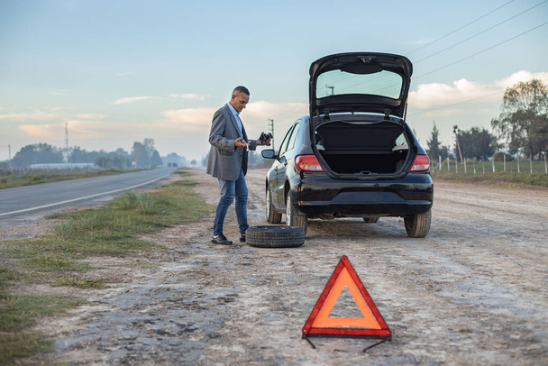 Confused latino man tries to change the flat tire of his car with copy space. - Fotografie, Obrázek