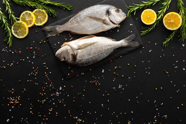 Dorada fish with lemon slices and rosemary sprigs on a slate board among spices on a black background, top view, copy space - Photo, Image