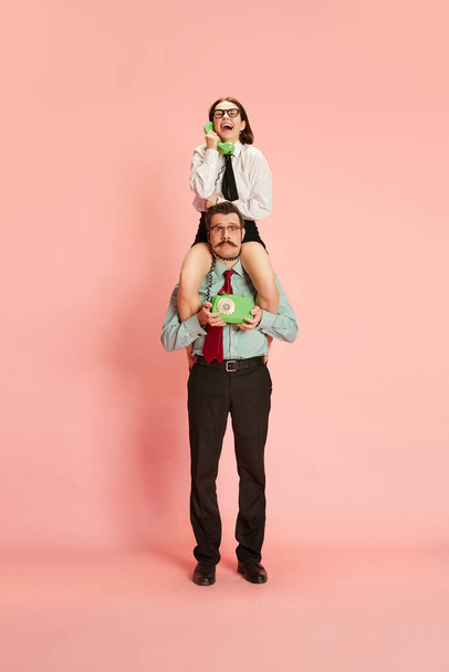 Portrait with office workers, shocked man and smiling woman sitting on male shoulders and speaking via telephone over pink studio background. Concept of business, career, relationship, emotions - Fotografie, Obrázek