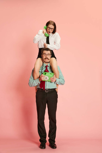 Business planning. Portrait with business couple man and shouting woman sitting on male shoulders and speaking via telephone over pink studio background. Concept of business, career, emotions - Foto, Bild