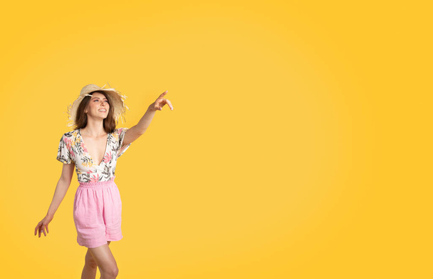 Letní sezónní nákupy. Okouzlující dáma v módním oblečení a slaměném klobouku, ukazující stranou na kopírovací prostor přes žluté pozadí studia. Obrovské slevy a propagační koncept - Fotografie, Obrázek