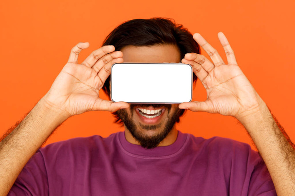 Eastern bearded smiling guy covering his eyes with horizontal smartphone with white empty screen, mockup on orange studio background. Social media, gadget addiction concept - Fotografie, Obrázek
