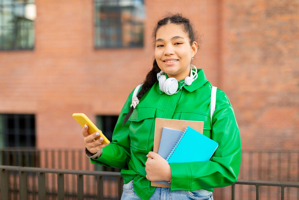 Portrait of happy mixed race student lady with smartphone and workbooks posing outdoors, female using mobile phone while standing outside near college - Фото, зображення