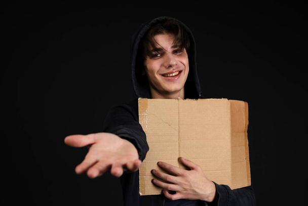 Улыбающийся бездомный молодой человек с синяками под глазами в грязной, рваной одежде, держащий пустую картонную вывеску и руку. Изолированный на черном фоне. - Фото, изображение