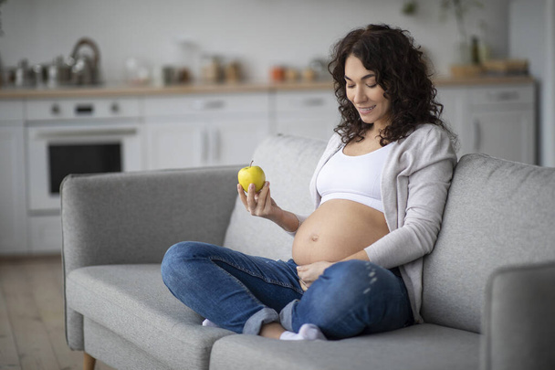 Healhy Snack. Young Pregnant Woman Eating Apple While Sitting On Couch At Home, Smiling Female Holding Fruit And Tenderly Touching Her Belly, Enjoying Vitamin Food During Pregnancy, Copy Space - 写真・画像